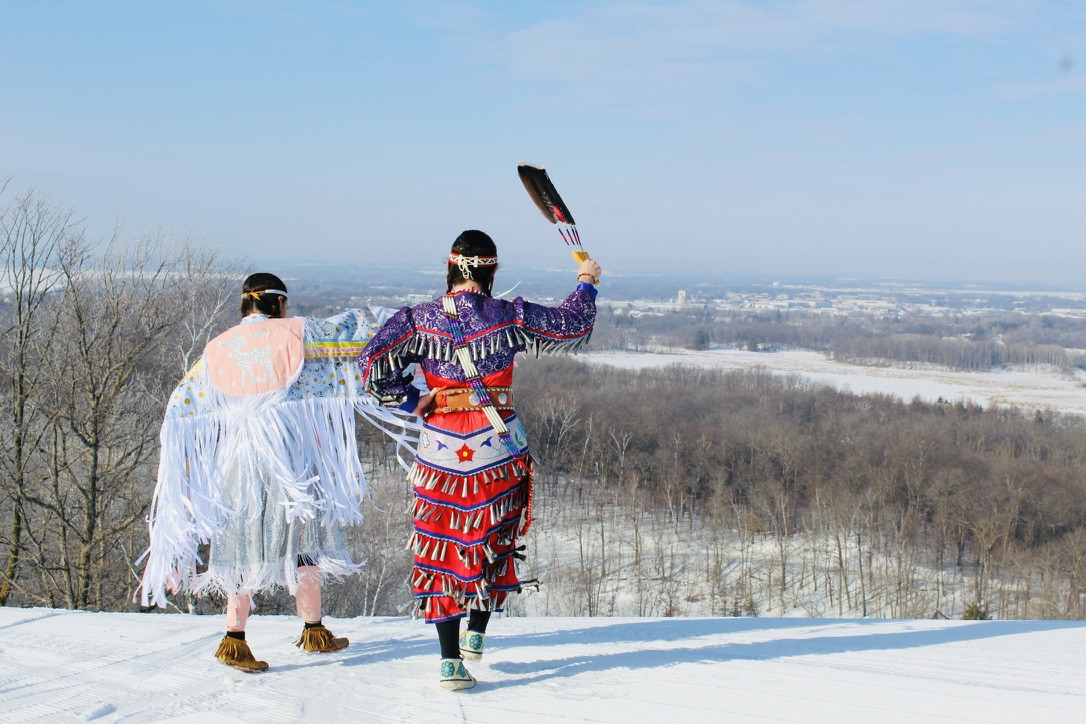 Two dancers in powwow regalia raising an eagle feather on a snowy ridge.